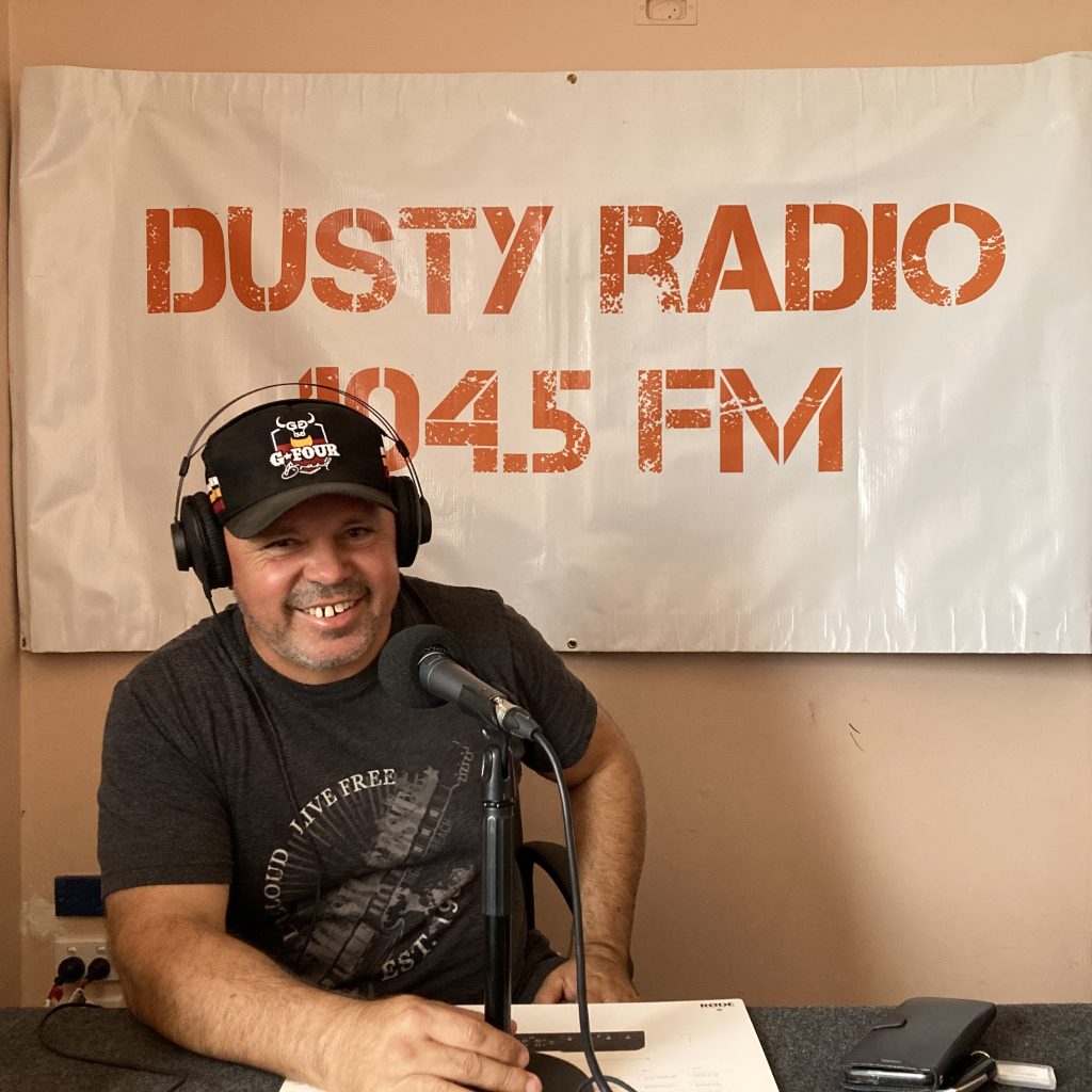 Local Coober Pedy man Jonathon Fatt-Clifton sits at a table behind a microphone, wearing headphones. Behind him is a banner that reads, "Dusty Radio 104.5 FM"